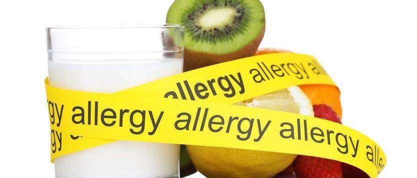 Ein Glas Milch mit verschiedenen Früchten und einer Schokoladentafel, umwickelt von einem gelben Band, das wiederholt 'Allergie' sagt.