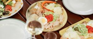 Auf einem Holztisch steht eine Pizza mit Fleisch, Käse und Gemüse, neben einem Glas Wein und Tellern.