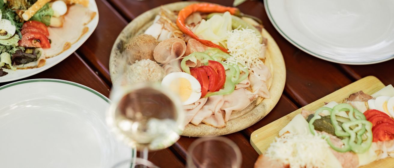 Auf einem Holztisch steht eine Pizza mit Fleisch, Käse und Gemüse, neben einem Glas Wein und Tellern.