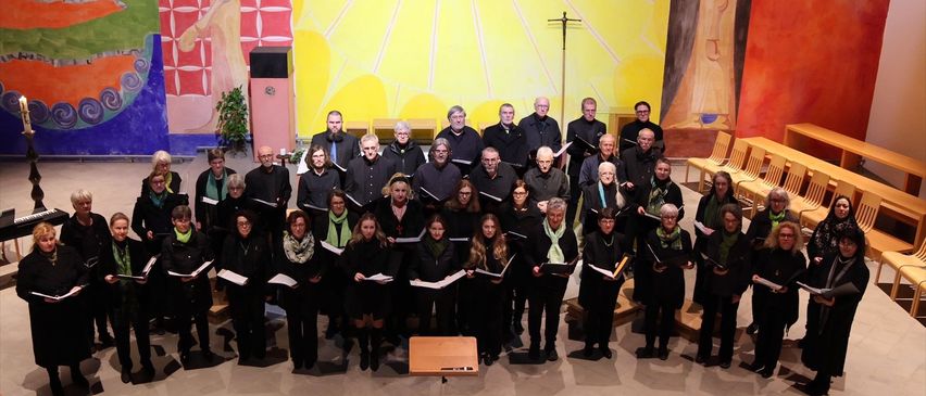 Ein Chor in schwarzen Kleidern steht auf der Bühne vor einem großen Wandbild. Der Chor hält ihre Notenblätter in den Händen und alle schauen in die gleiche Richtung. Dahinter befindet sich ein hölzerner Rednerpult.
