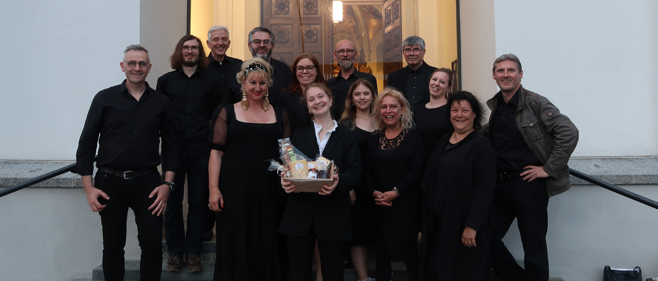 Eine Gruppe von Menschen in Schwarz posiert für ein Foto auf den Stufen eines Gebäudes. Eine Frau in der Mitte hält einen Korb mit Geschenken. Dahinter befindet sich eine Glaswand mit einer Tür und einer Lampe.