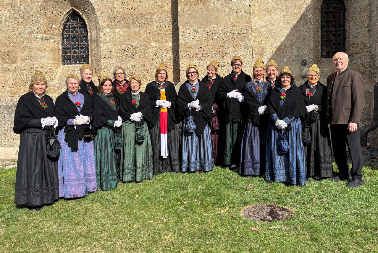 Eine Gruppe von Frauen in traditioneller Kleidung mit weißen Handschuhen und Hüten posiert für ein Foto vor einem Steingebäude mit einem Fenster.