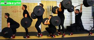 Fünf Männer in schwarzen Trainingsklamotten führen Kniebeugen mit Hanteln in einem Fitnessstudio durch. Sie halten alle Hanteln über ihren Köpfen. Sie stehen in einer Reihe. Die Wand hinter ihnen besteht aus Holz und hat ein weißes Fliesendesign.