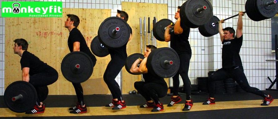 Fünf Männer in schwarzen Trainingsklamotten führen Kniebeugen mit Hanteln in einem Fitnessstudio durch. Sie halten alle Hanteln über ihren Köpfen. Sie stehen in einer Reihe. Die Wand hinter ihnen besteht aus Holz und hat ein weißes Fliesendesign.