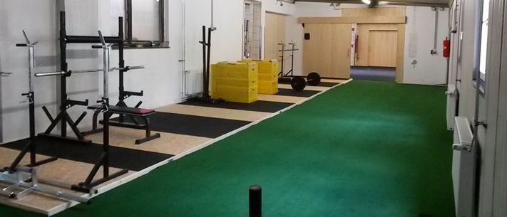 Ein leerer Fitnessraum mit grünem Teppich, verschiedenen Trainingsgeräten, gelben Lagerboxen und weißen Wänden.