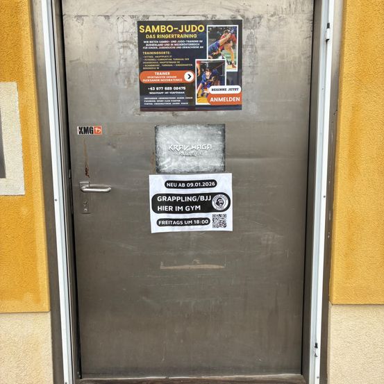 Eine Metalltür mit einer gelben Wand. Sie hat ein Schild, das SAMBO-JUD liest, und ein kleineres Schild, das Grappling/BJJ hier im Fitnessstudio liest. Freitags um 18:00.