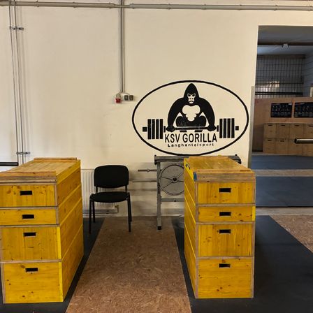 Ein Fitnessstudio mit einer weißen Wand, auf der ein Gorilla-Logo und gelbe Holzschränke auf beiden Seiten eines Stuhls zu sehen sind. Der Fitnessraum hat einen schwarzen Teppich auf dem Boden.
