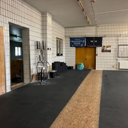 Ein leerer Gewichtheberraum mit einem zentralen Pfad und Bannern, die 'Dies ist ein Gewichtheberhaus' und 'WH' lesen. Ausrüstung und eine Tasche liegen auf dem Boden.