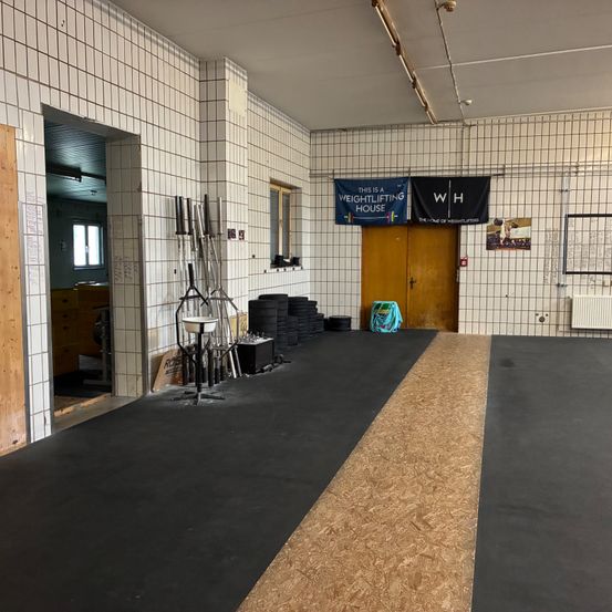 Ein leerer Gewichtheberraum mit einem zentralen Pfad und Bannern, die 'Dies ist ein Gewichtheberhaus' und 'WH' lesen. Ausrüstung und eine Tasche liegen auf dem Boden.