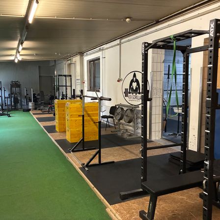 Ein Fitnessstudio mit grünem Boden, ausgestattet mit verschiedenen Trainingsgeräten wie einem Kniebeugenständer, gelben Gewichtsboxen und einer Bank. Es gibt auch mehrere Stühle und einen Schreibtisch mit einem schwarzen runden Logo.