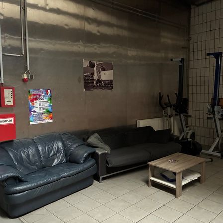 Ein Fitnessraum mit gefliestem Boden und grauen Wänden. Zwei Ledersofas stehen an einer Wand mit Fitnessgeräten in der Nähe. Ein hölzerner Tisch mit einem Kissen steht in der Mitte. Poster sind an der Wand befestigt.