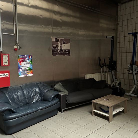 Ein Fitnessraum mit gefliestem Boden und grauen Wänden. Zwei Ledersofas stehen an einer Wand mit Fitnessgeräten in der Nähe. Ein hölzerner Tisch mit einem Kissen steht in der Mitte. Poster sind an der Wand befestigt.