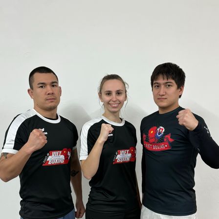 Drei Personen in schwarzen Viet Boxing-Shirts stehen zusammen. Die beiden Männer haben ihre Fäuste erhoben, und die Frau lächelt.