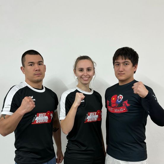 Drei Personen in schwarzen Viet Boxing-Shirts stehen zusammen. Die beiden Männer haben ihre Fäuste erhoben, und die Frau lächelt.