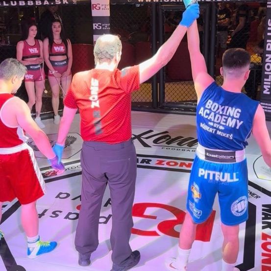 Ein Schiedsrichter in einem roten Shirt und Handschuhen hebt die Hand eines siegreichen jungen Boxers in einem blauen Outfit hoch, während zwei weitere junge Boxer in roten Uniformen daneben stehen.