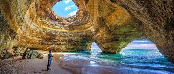Eine Person steht auf einem sandigen Strand in der Nähe einer Höhle mit einer großen Öffnung zum Meer. Das Sonnenlicht scheint durch das Loch in der Höhle.