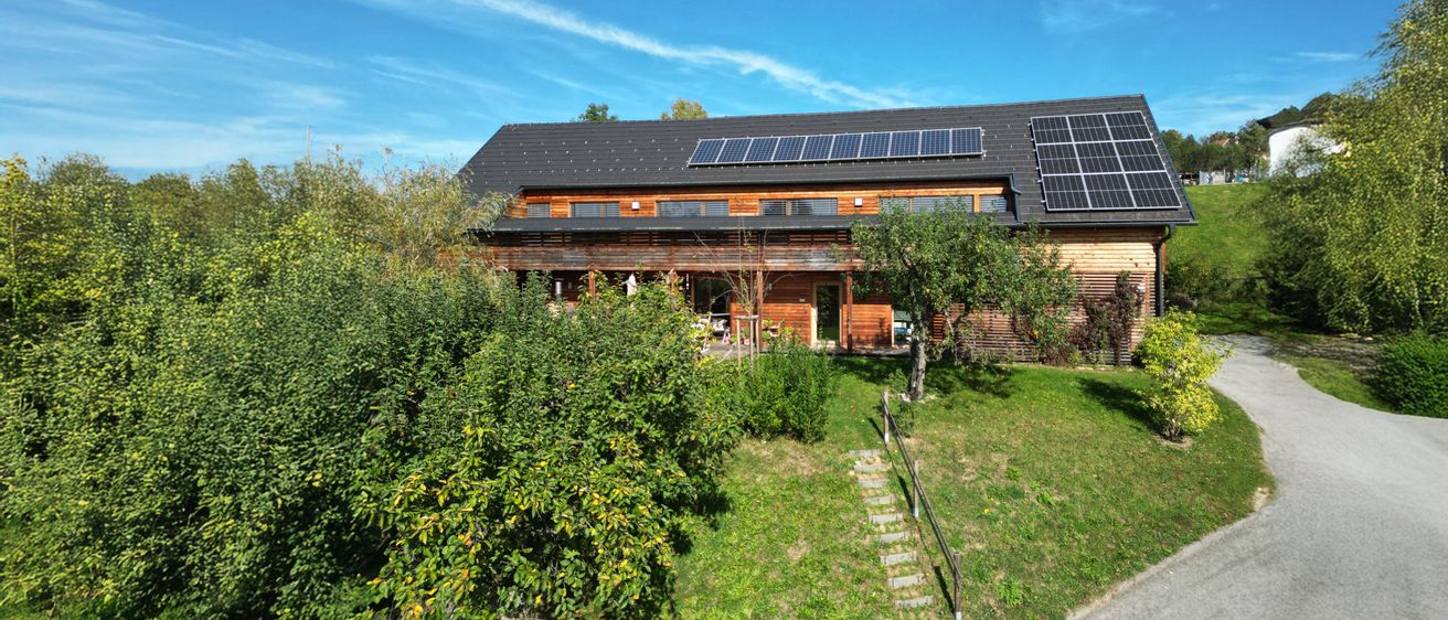 Ein Holzhaus mit schwarzem Dach, Solarmodulen und einem Garten mit Bäumen und Büschen an einem sonnigen Tag.
