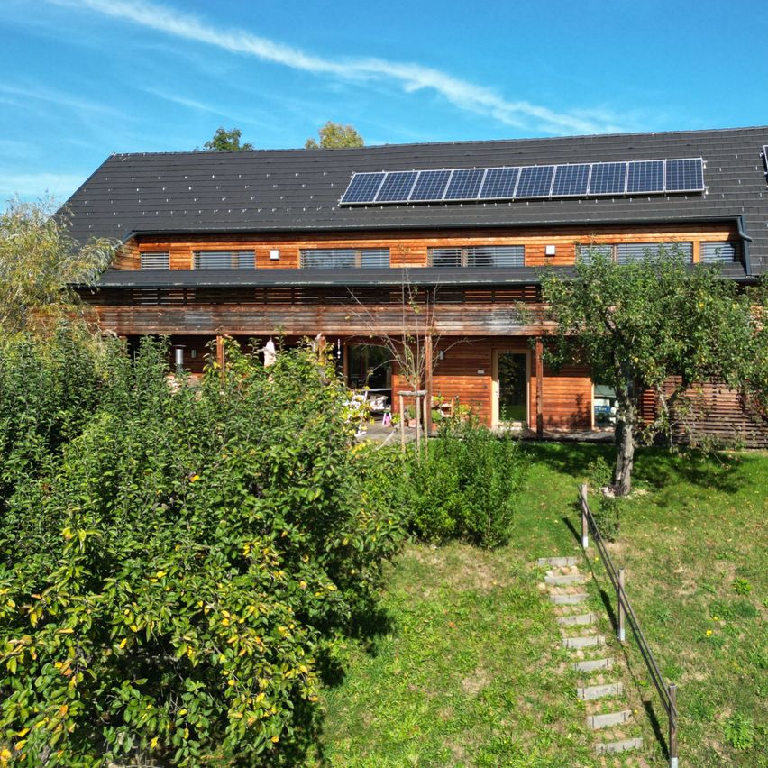 Ein Holzhaus mit schwarzem Dach, Solarmodulen und einem Garten mit Bäumen und Büschen an einem sonnigen Tag.