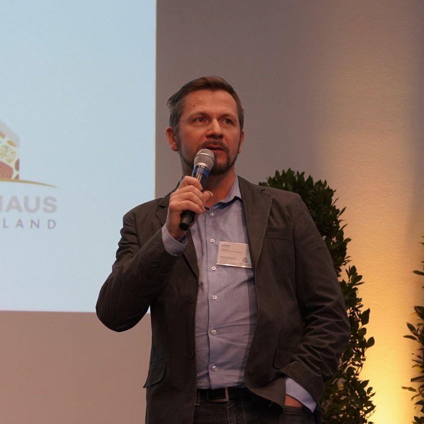Ein Mann in einem Anzug spricht in ein Mikrofon, hält es mit der rechten Hand und steht vor einer Projektionsleinwand.