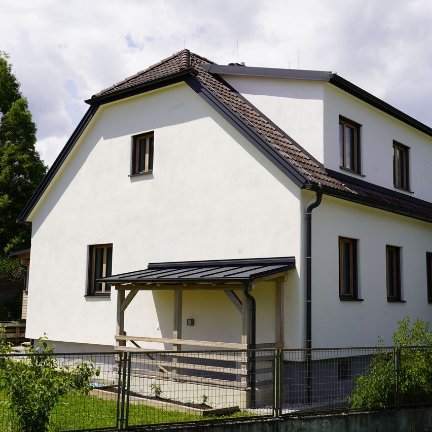 Ein weißes Haus mit einem braunen Dach und schwarzen Abschlüssen. Es hat einen kleinen Vorbau mit schwarzem Dach, mehreren Fenstern und einem Zaun davor.