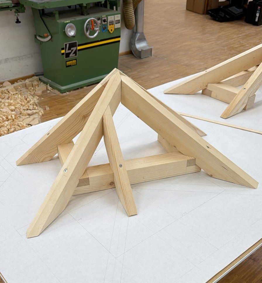 Eine Holzbinderstruktur wird auf einer weißen Oberfläche zusammengesetzt. In der Nähe sind eine grüne Maschine und ein Haufen Holzspäne zu sehen.