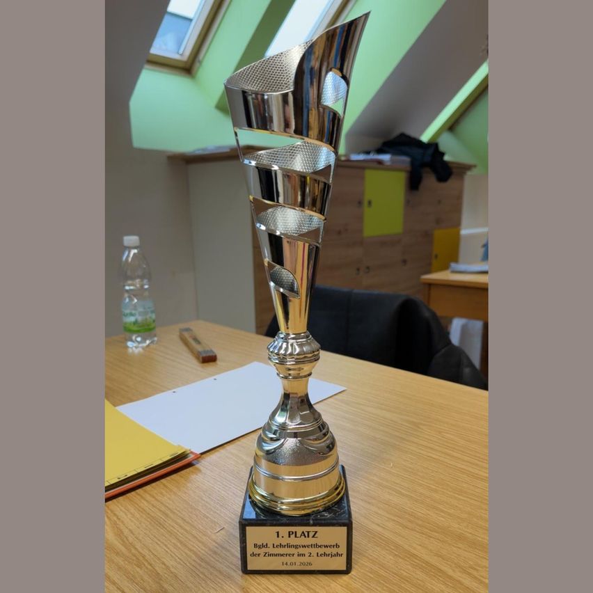 Ein silberner Pokal auf einem Holztisch, beschriftet mit '1. Platz' für das Jahr 2026, mit einer durchsichtigen Wasserflasche und Papieren im Hintergrund.