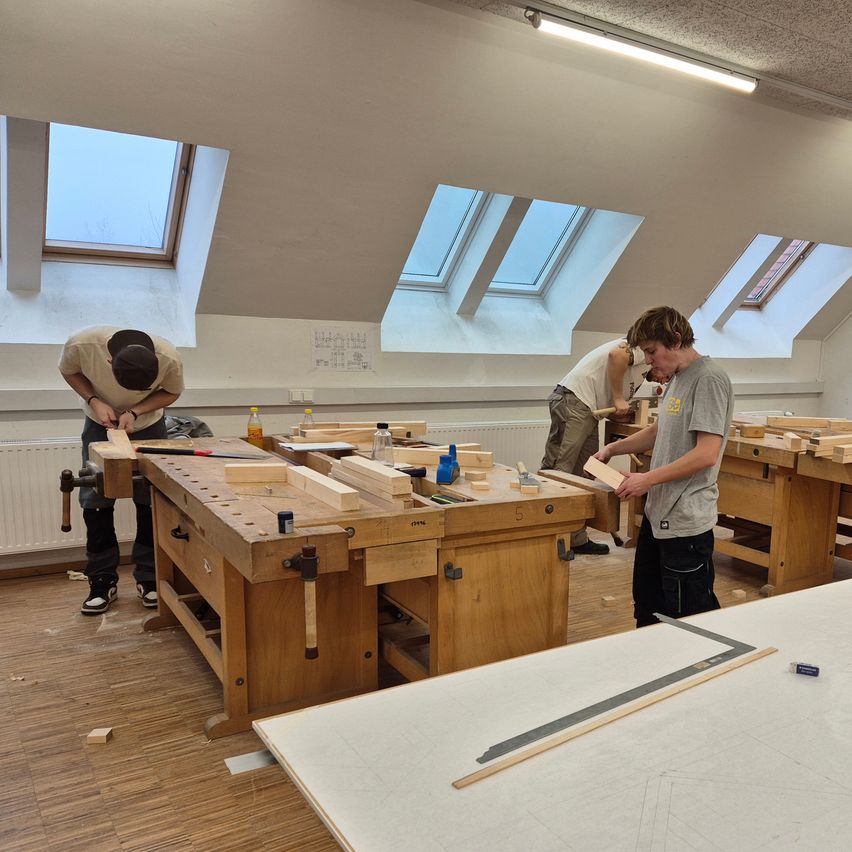 Drei Personen arbeiten in einer Werkstatt mit Holzwerkbänken, umgeben von verschiedenen Werkzeugen. Sie sind mit Zimmermannsarbeiten beschäftigt.