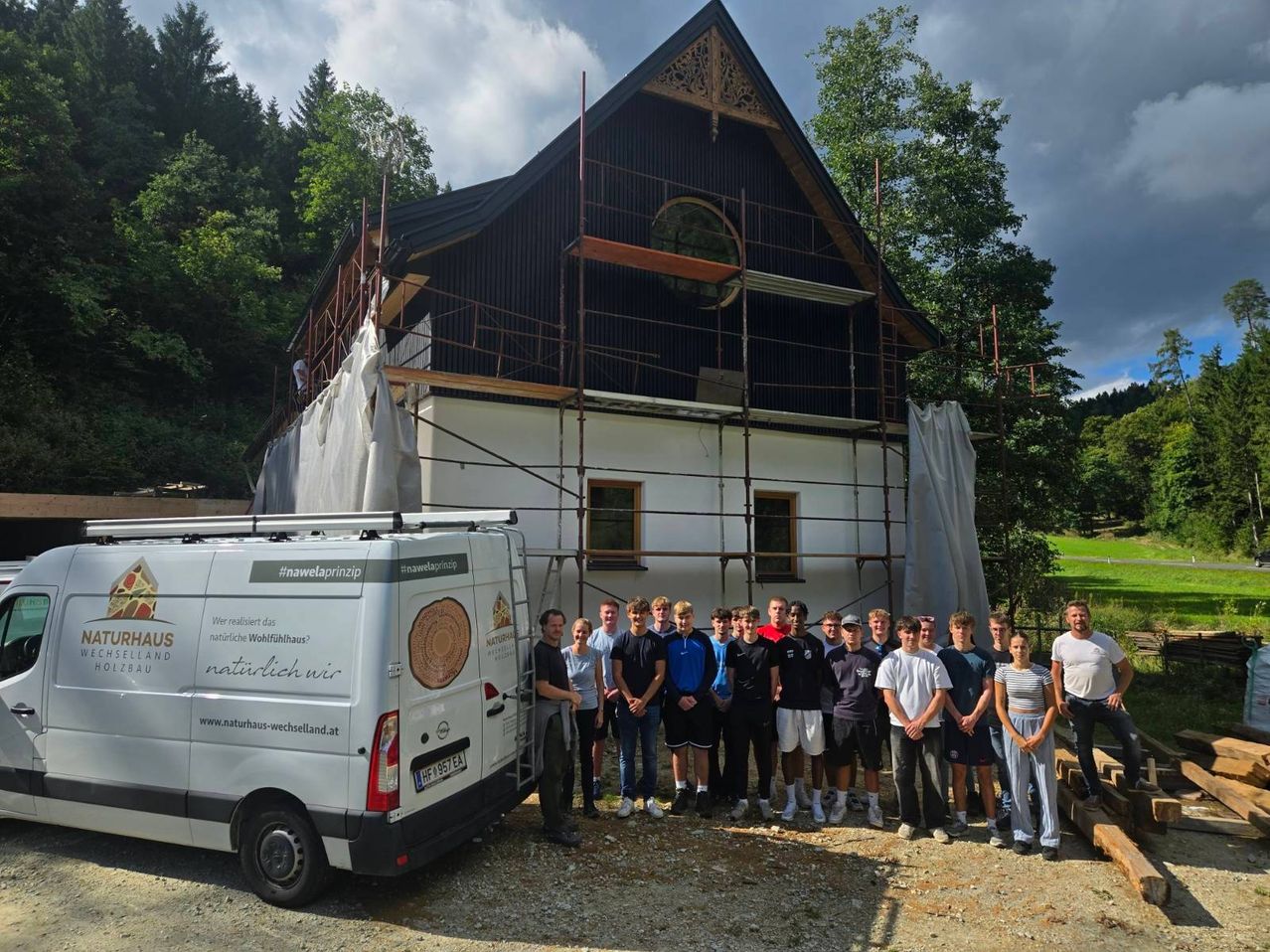 A group of individuals stands in front of a building under renovation. The building has scaffolding, and a van is parked nearby.