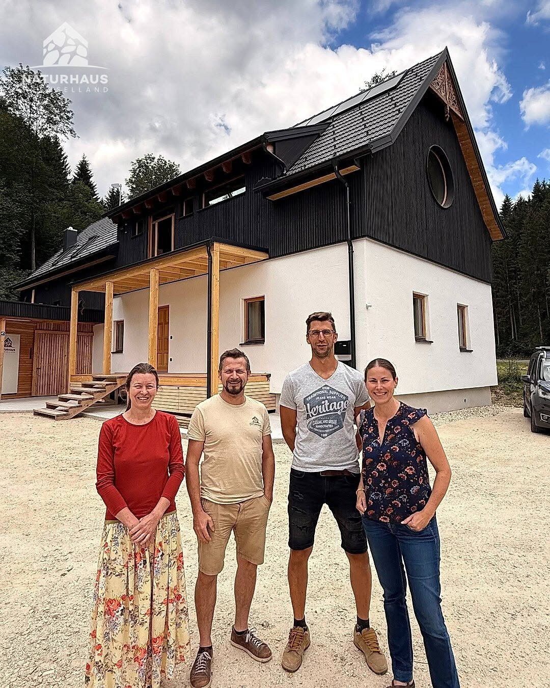 Four people stand in front of a modern house with a black and white exterior, solar panels on the roof, and a circular window. They are smiling and appear to be posing for a photo.