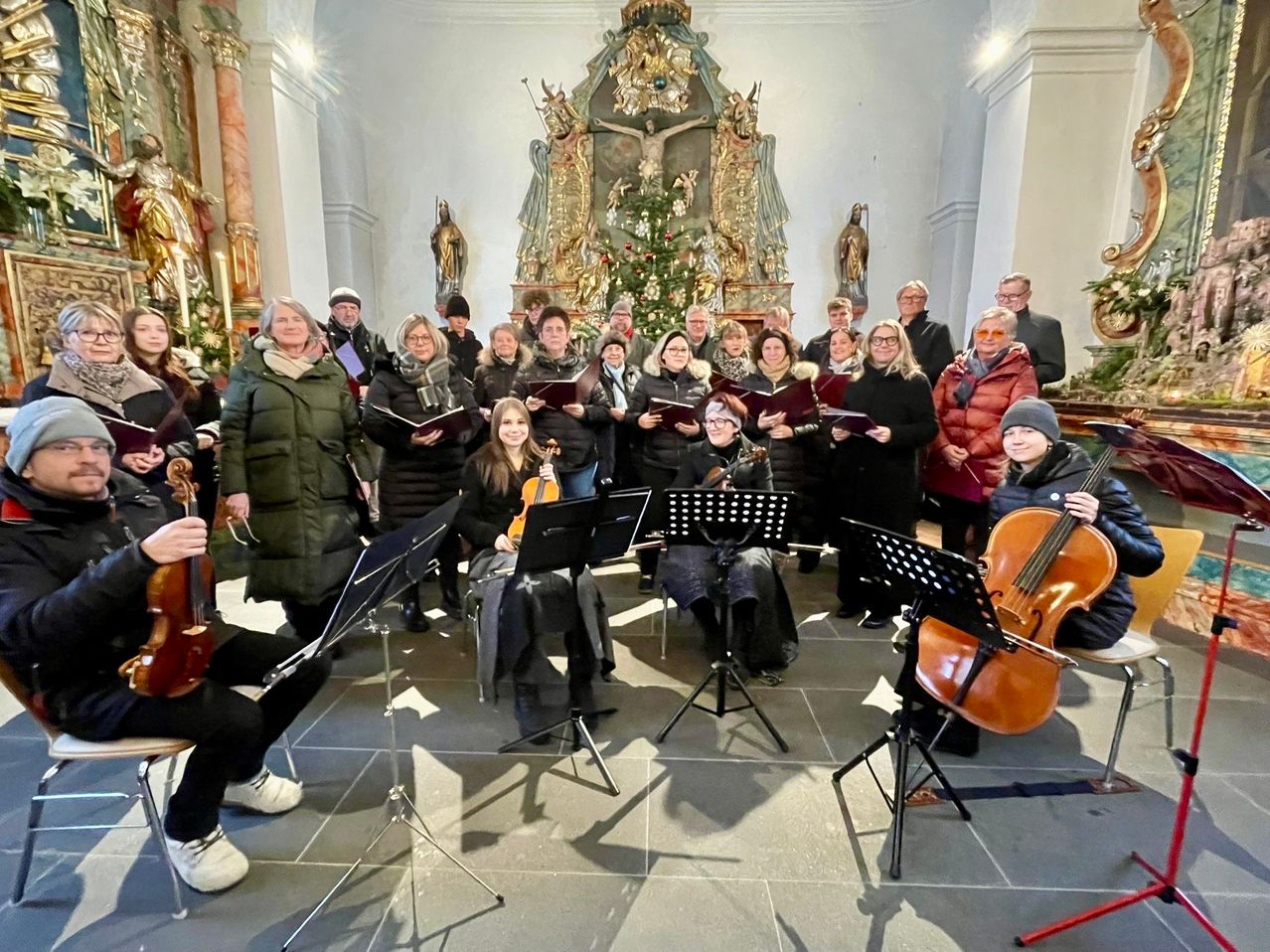 Eine Gruppe von Menschen in Winterkleidung versammelt sich in einer Kirche, spielt Violinen und Celli, mit Notenständern vor ihnen.