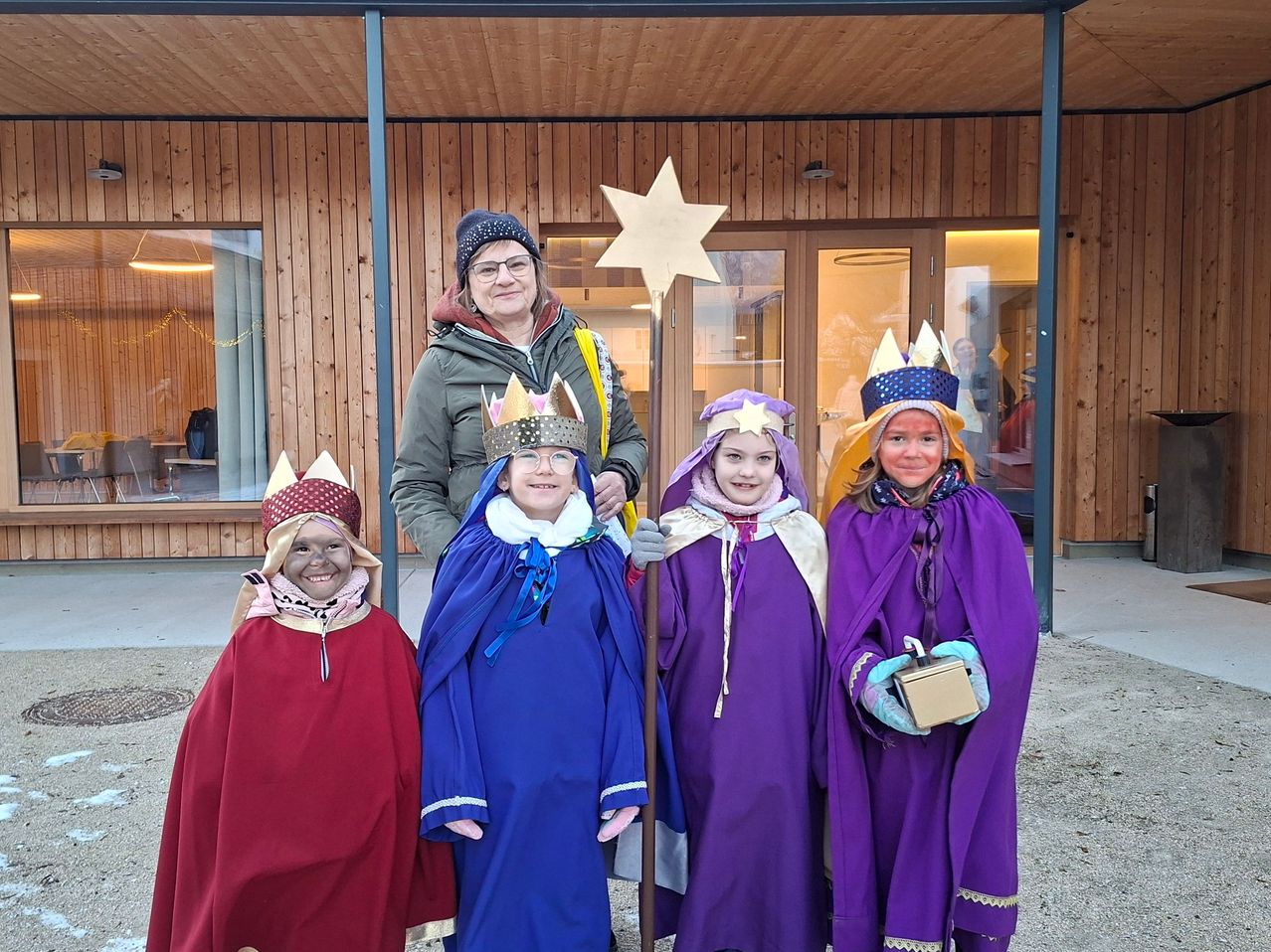 Eine Frau mit einem Stern und vier Kinder in mittelalterlichen Kostümen stehen vor einem Holzgebäude. Die Kinder tragen Kronen und halten Geschenke.