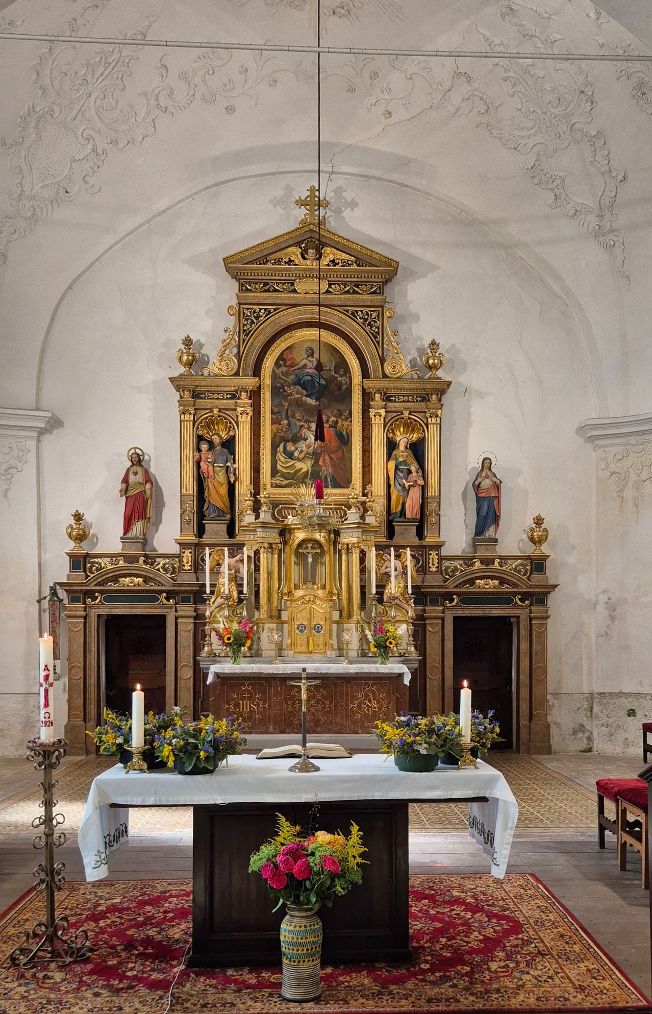 An ornate altar in a church with a central painting, flanked by statues, and adorned with candles and flowers.