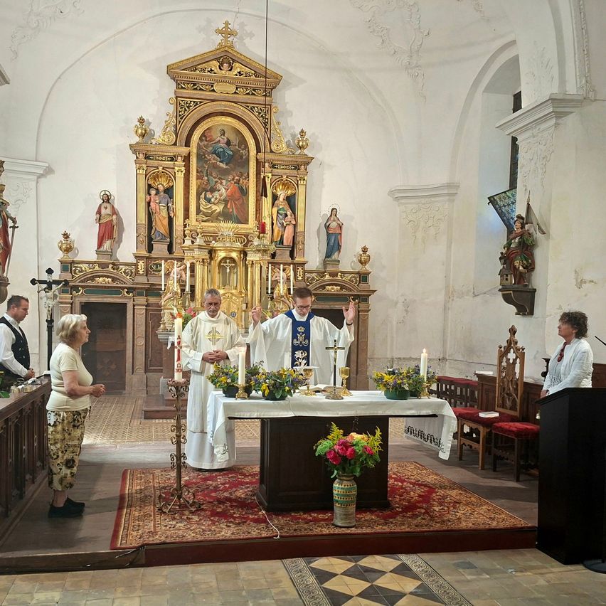 Two priests stand at an altar in a church, surrounded by candles and flowers, with a woman watching from the side.