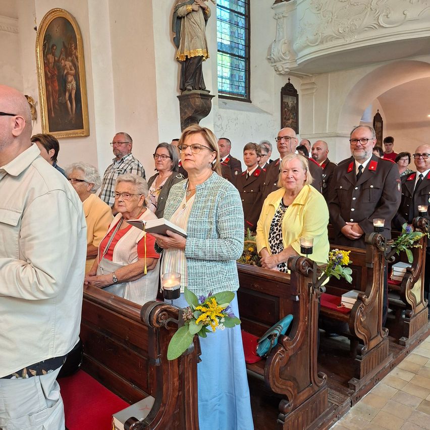 A group of people, some in formal attire, stand in a church during a service, holding candles and books.