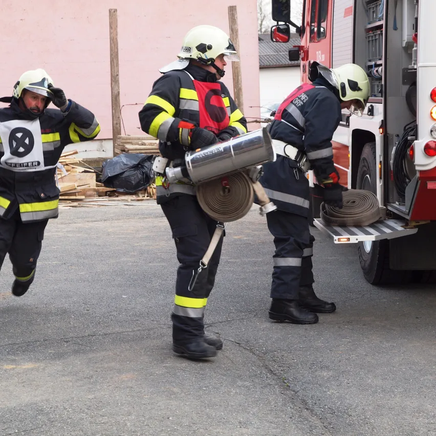 Drei Feuerwehrleute sind draußen. Einer hält eine Feuerwehrschläuche, während ein anderer an einem Fahrzeug lehnt. Der dritte geht auf das Fahrzeug zu.