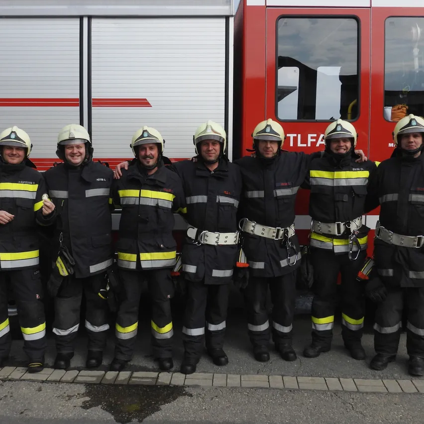Sechs Feuerwehrleute stehen vor einem roten Feuerwehrauto. Sie tragen alle einheitliche Uniformen und Helme.
