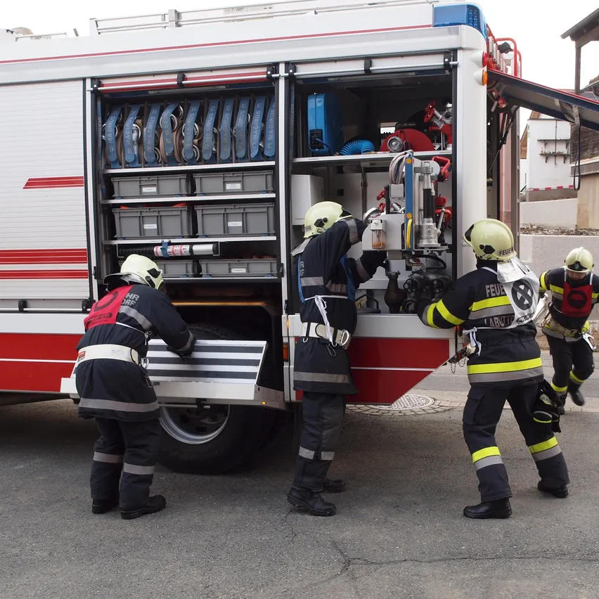 Feuerwehrleute versammeln sich um einen offenen Feuerwehrwagen und bereiten Ausrüstung vor. Ein Feuerwehrmann hält ein Werkzeug in der Hand.