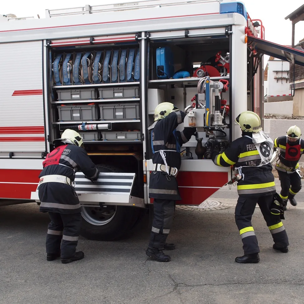 Feuerwehrleute versammeln sich um einen offenen Feuerwehrwagen und bereiten Ausrüstung vor. Ein Feuerwehrmann hält ein Werkzeug in der Hand.