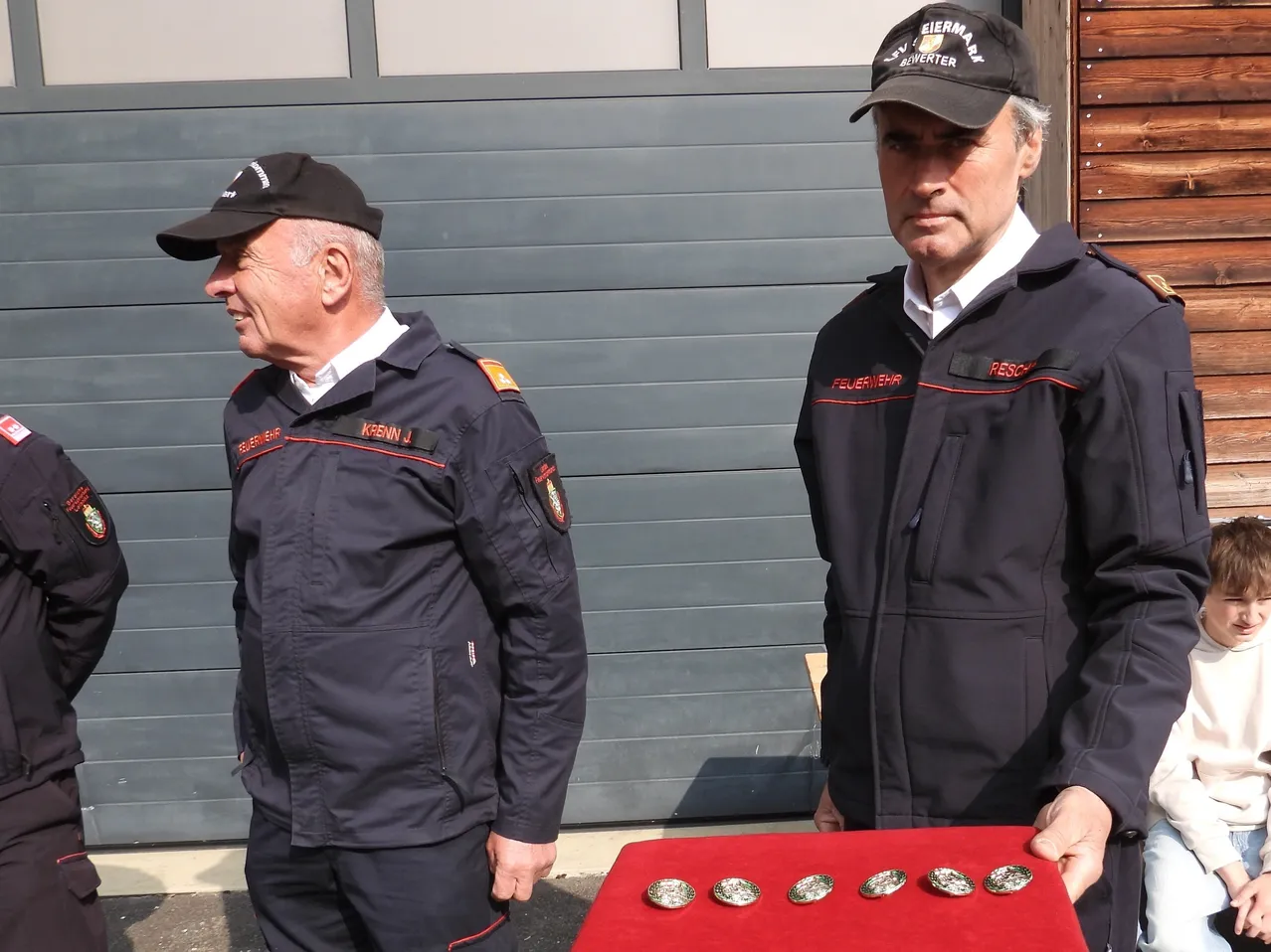 Zwei Feuerwehrleute stehen neben einer roten Ausstellung mit Medaillen, einer trägt einen Hut mit 'Eiermarkt' und der andere mit 'Feuerwehr' darauf.