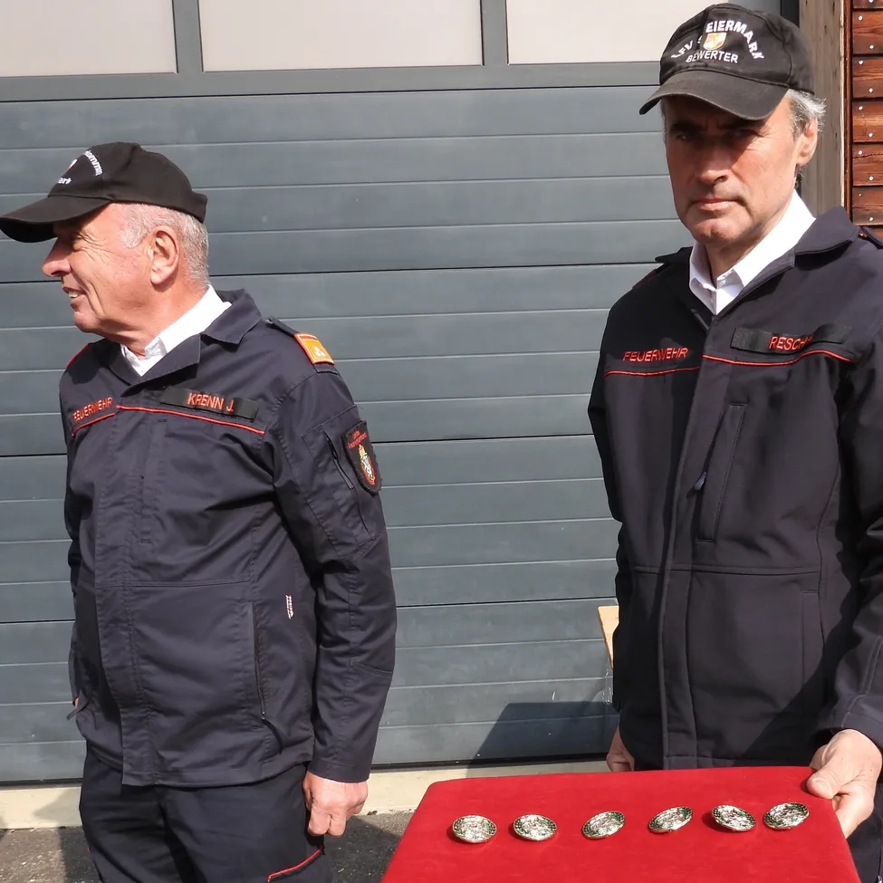 Zwei Feuerwehrleute stehen neben einer roten Ausstellung mit Medaillen, einer trägt einen Hut mit 'Eiermarkt' und der andere mit 'Feuerwehr' darauf.