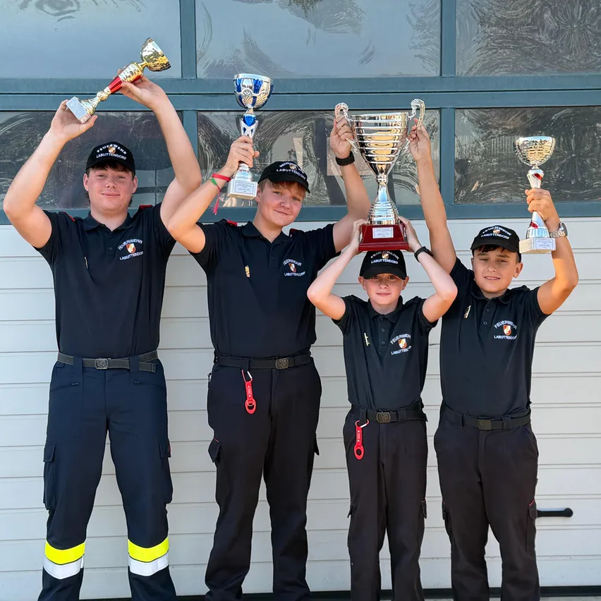 Vier junge Männer in identischen Uniformen posieren mit ihren Trophäen. Sie stehen vor einer weißen Garagentür.