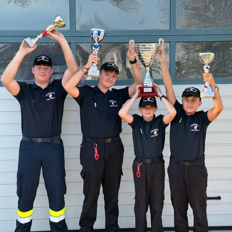 Vier junge Männer in identischen Uniformen posieren mit ihren Trophäen. Sie stehen vor einer weißen Garagentür.
