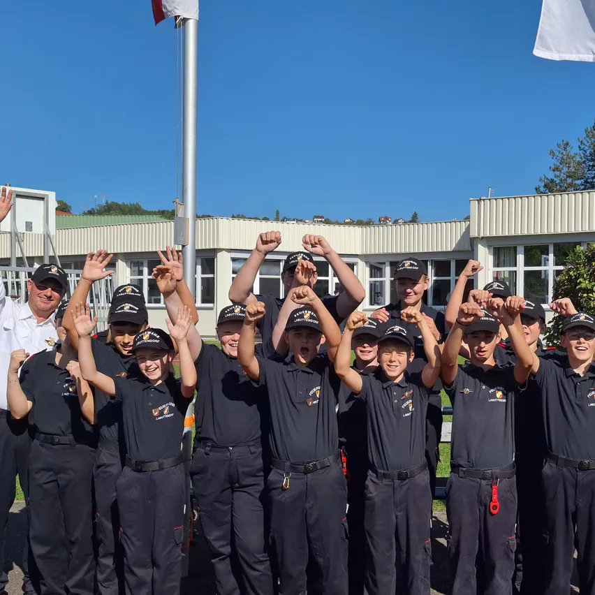 Eine Gruppe von Menschen in schwarzen Uniformen, wahrscheinlich ein Sportteam, posiert für ein Foto mit erhobenen Händen vor einem Gebäude.
