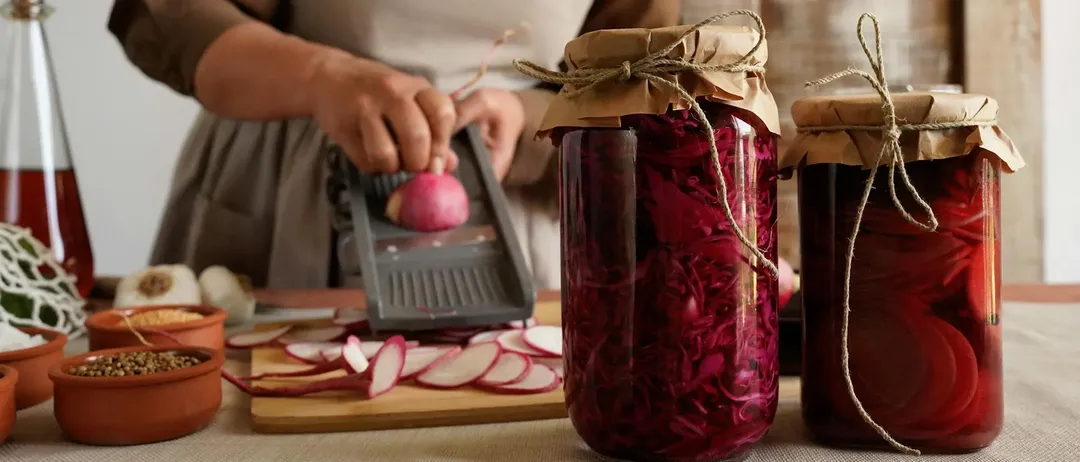Eine Person in einer Schürze schneidet einen roten Rettich mit einem Gemüseschneider. Vor ihr stehen zwei Gläser mit eingelegten roten Rettichen mit braunen Papierdeckeln.
