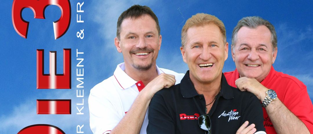 Drei Männer stehen lächelnd vor einem blauen Himmelshintergrund mit dem Wort 'Die' in roten Buchstaben und dem Text 'Korner Klemens & Frei' auf der linken Seite. Der Mann in der Mitte trägt ein schwarzes Shirt mit dem Text 'Austrian Wear' und einer Uhr. Die anderen beiden Männer tragen rote und weiße Hemden.