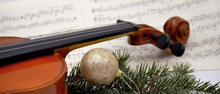 Eine Geige ruht auf einem Musikbogen mit Tannenzweigen und einer goldenen Weihnachtskugel im Vordergrund. Die Musiknoten sind teilweise verschwommen.
