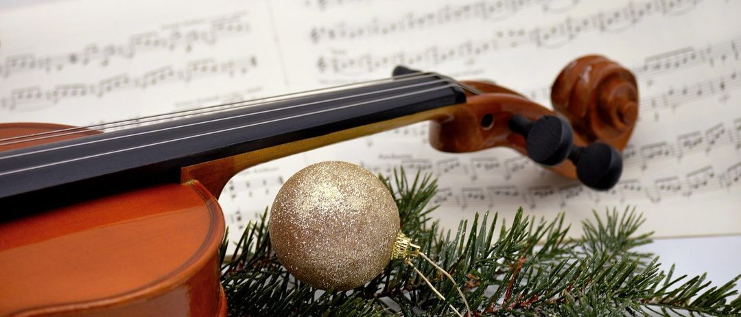 Eine Geige ruht auf einem Musikbogen mit Tannenzweigen und einer goldenen Weihnachtskugel im Vordergrund. Die Musiknoten sind teilweise verschwommen.