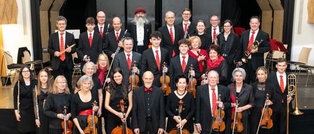 Eine Gruppe von Musikern in formeller Kleidung posiert für ein Foto. Einige halten ihre Instrumente. Ein Mann in der Mitte trägt eine rote Fliege. Der Hintergrund ist unscharf.