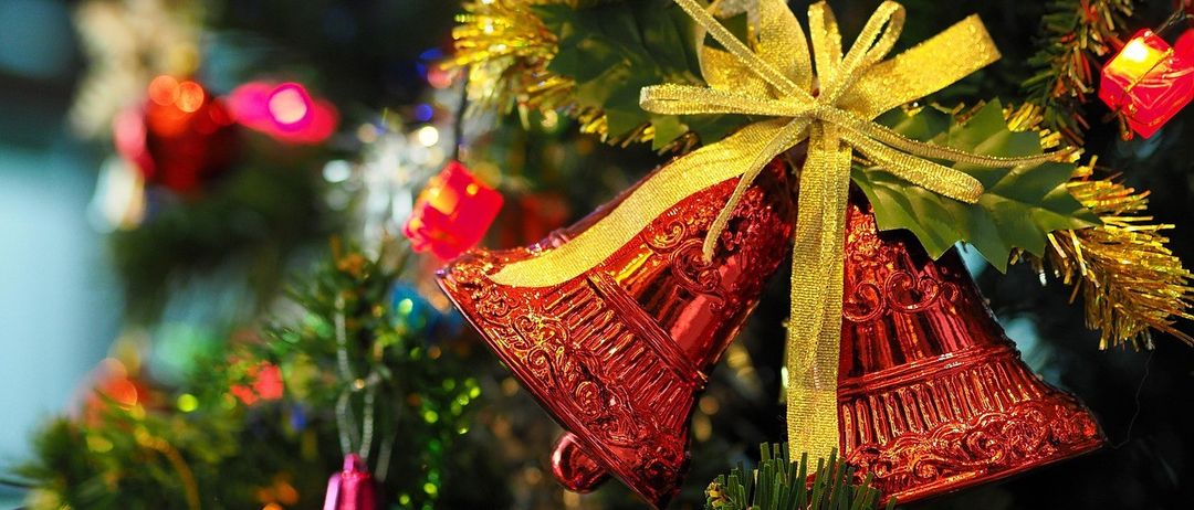 Nahaufnahme von roten Weihnachtsglocken mit einem goldenen Band, die an einem Baum hängen, umgeben von bunten Ornamenten und Lichtern.