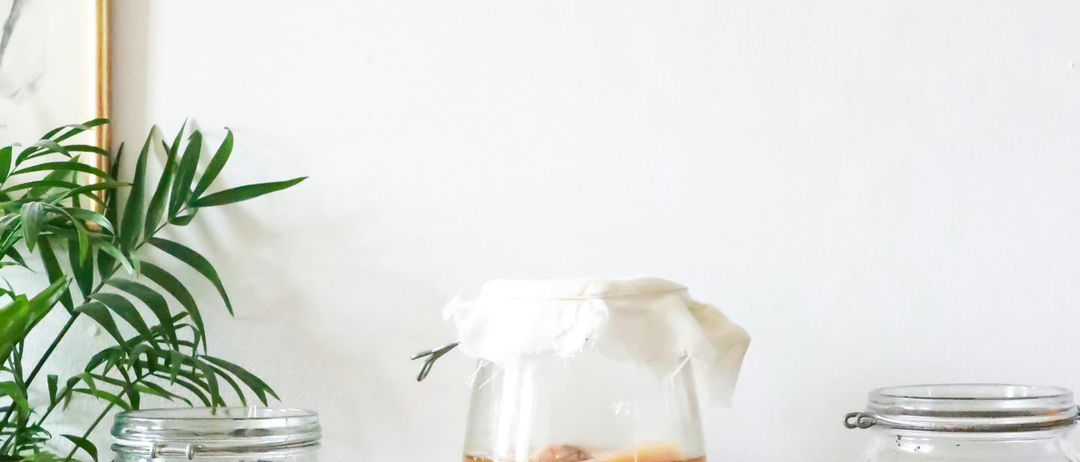 Drei Glasflaschen mit brauner Flüssigkeit und weißen Tüchern stehen auf einem Tisch an einer weißen Wand, darüber eine Kupferuhr.